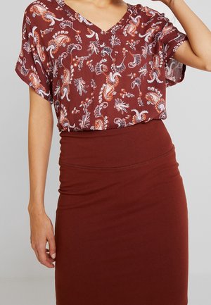 Woman wearing a maroon floral-patterned blouse with short sleeves and a plain maroon fitted skirt against a gray background.