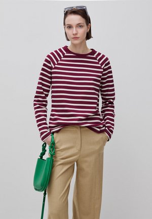 Młody dorosły z krótkimi brązowymi włosami, ubrany w bordowo-białą koszulę w paski z długim rękawem, beżowe spodnie, trzyma zieloną torebkę i ma okulary przeciwsłoneczne na głowie.