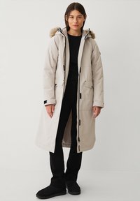 Parka lunga beige con cappuccio foderato in pelliccia, chiusura frontale con zip e bottoni a pressione, multiple tasche e polsini regolabili. Indossata sopra un outfit nero.