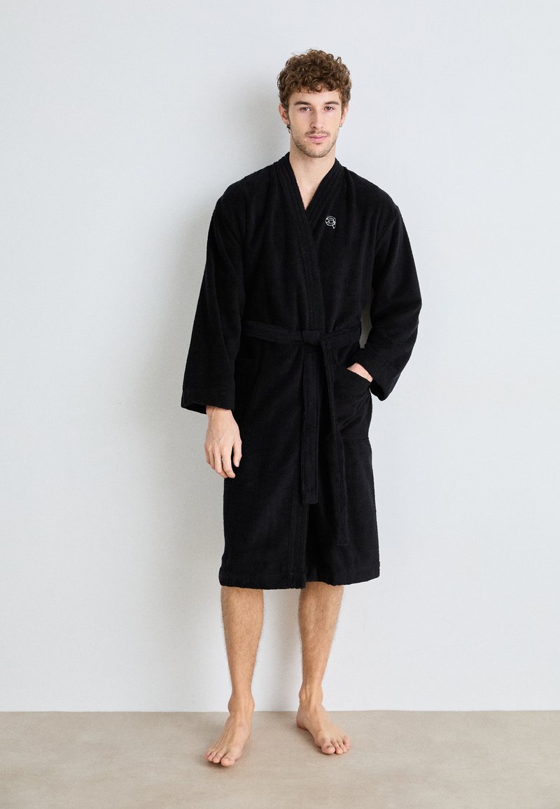 Roupa de banho preta em tecido felpudo, com mangas longas, cinto atado e bolsos laterais, apresentando um pequeno logo bordado no peito.