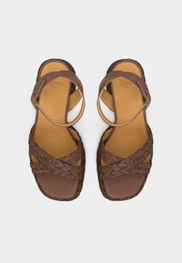 Καφέ σανδάλια με παχιά πλεκτά λουριά που  διασταυρώνονται πάνω από τα δάχτυλα και λουράκια αστραγάλου με αγκράφες, επίπεδη σόλα και σχεδιασμό με τετράγωνη μύτη.