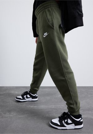 Person iført olivengrønne Nike træningsbukser og sort-hvide Nike sneakers, der står på gråt gulv med en ensfarvet lys baggrund.