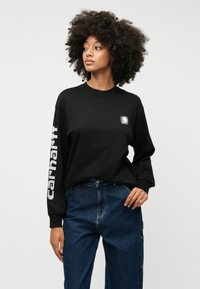 Melnas garās piedurknes krekliņš ar mazu logotipu uz krūtīm un spilgtiem baltajiem burtiem uz kreisās piedurknes. Savienots ar ziliem džinsa biksēm.