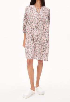 Frau trägt ein lockeres, knielanges Kleid mit Blumenmuster, dreiviertel Ärmeln und weißen Slip-On-Clogs, steht vor einem einfarbigen Hintergrund.