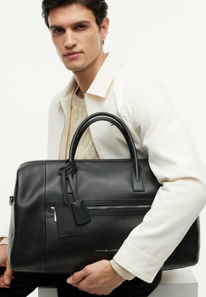 Jeune homme en veste blanche tenant une grande mallette noire en cuir Tommy Hilfiger avec deux poignées et une poche zippée à l'avant.