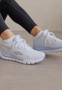 Fehér sportcipők hálós felsőrésszel, világoskék díszítéssel és texturált talppal. A fűzők szépen meg vannak kötve. Fekete leggings és fehér zoknik kíséretében viselve.