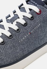 Sinine denimist kets, millel on valged paelad, kummitald, mustad silmad ja "TOMMY HILFIGER" logo. Omab tekstuuriga kangast pealistust.