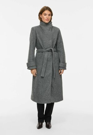 Femme portant un long manteau en laine gris ceinturé avec un col montant, pantalon noir et chaussures foncées, debout sur un fond blanc uni.