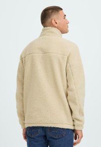 Lys beige fleecejakke med høj krave, tekstureret overflade og løs pasform. Bæres med blå jeans, der viser et simpelt design uden synlige logoer.