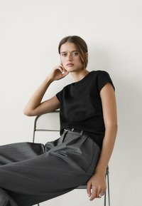 Černý top s krátkým rukávem a volným střihem, kombinovaný s šedými kalhotami na míru s skladovým designem a detailem opasku.