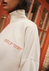 Kreemist särgist, millel on kõrge kaelus ja luku sulgemine, on brodeeritud oranž kiri: "SELF ATHLEISURE EST. 2023." Pehme kangas, mugav lõige, minimalistlik disain.