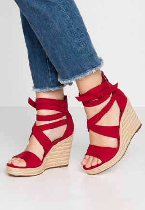 Røde ruskind wedge sandaler med krydset remme og ankelbindinger, med en struktureret naturlig reb-wedge hæl og åbne tæer. Denim cropped bukser er synlige.