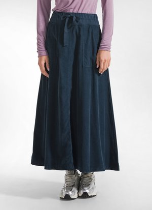 Persoon draagt een lange marineblauwe rok met voorzakken, een roze top met lange mouwen en zilveren sportschoenen, staand op een lichte achtergrond.