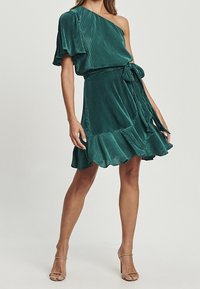 Robe verte à manches courtes et à une épaule, fabriquée en tissu plissé, avec un lien à la taille et une jupe évasée avec un ourlet volanté.