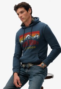 Felpa blu con grafica di montagne, cappuccio con cordino e tasca a marsupio. Indossata sopra una camicia di flanella a quadri, abbinata a jeans blu.