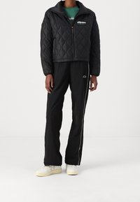 Czarna pikowana kurtka z wysokim kołnierzem, zamek z przodu i detalami logo, w połączeniu z czarnymi spodniami z bocznymi paskami i jasnymi sneakerami.