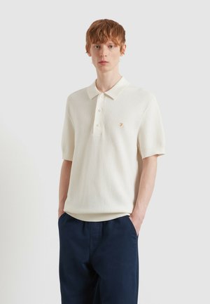 Junger Mann mit roten Haaren trägt ein weißes, kurzärmeliges Poloshirt mit Struktur und dunkelblaue Hosen, steht mit den Händen in den Taschen.