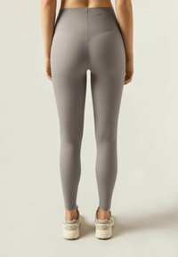 Jasnoszare legginsy z wysokim stanem, wykonane z elastycznego materiału, charakteryzujące się bezszwowym designem i minimalnym brandingiem. W zestawie z beżowymi butami sportowymi.