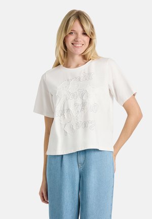 Lächelnde Frau mit blondem Haar trägt ein weißes kurzärmliges Shirt mit Blumenmuster und hellblaue High-Waist-Jeans vor einfarbigem Hintergrund.