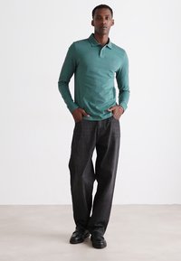 Cămașă polo cu mânecă lungă, de culoare teal, cu un guler clasic, combinată cu pantaloni negri din denim largi și pantofi negri, pe un fundal neutru.