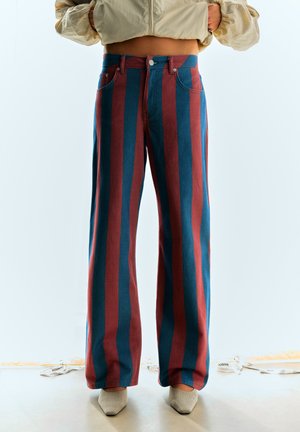 Personne portant un pantalon taille haute à jambes larges avec des rayures verticales bleues et rouges, assorti à des chaussures beiges à bouts pointus et une veste courte légère.