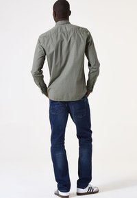 Groen gemusterd shirt met lange mouwen, knoopmanchetten en een afgeronde zoom; gedragen met een blauwe spijkerbroek en witte sneakers met zwarte strepen.