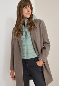 Brauner Wollmischung langer Mantel über einer hellgrünen gesteppte Daunenjacke. Die Daunenjacke hat einen Reißverschluss vorne und zeichnet sich durch ihre strukturierte Oberfläche aus.