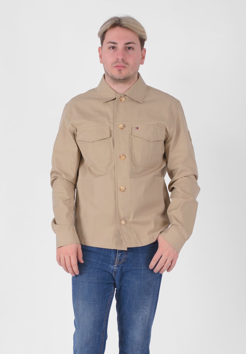 Giacca khaki con bottoni, due tasche sul petto, bottoni in legno naturale e colletto, abbinata a jeans blu su uno sfondo semplice.