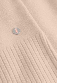 Licht beige stof met een horizontale ribbelstructuur en een kleine zilveren ronde bedel met een roze hondensilhouet.