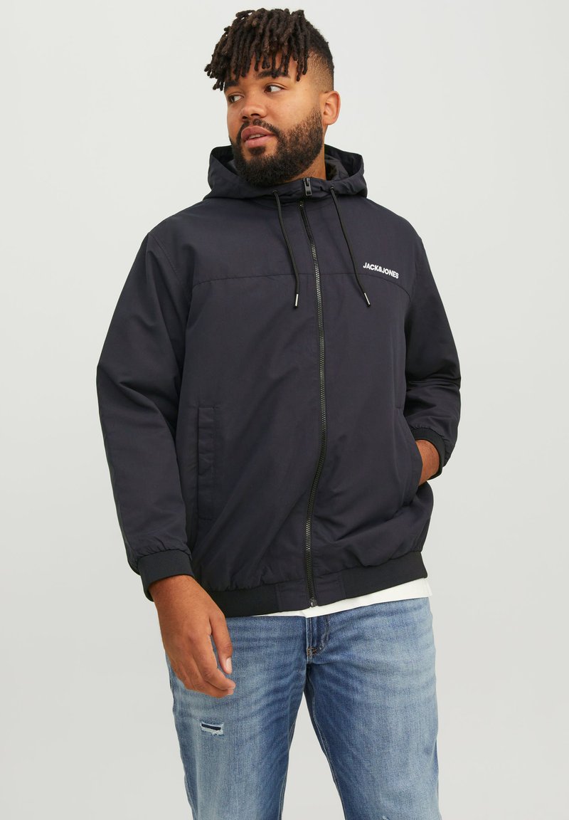 Navy blue hooded jacket with elastic cuffs, zip front, and logo on the chest. Worn over a white shirt, paired with distressed light blue jeans.