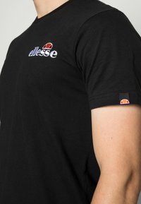 Närbild av en person med en svart T-shirt med "ellesse"-logotyp broderad på bröstet och en liten logotag på ärmen.