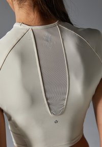 Light beige athletic top featuring a mesh panel at the back, structured seams, and a small logo at the center lower front.