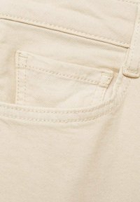 Beige denim fabric close-up featuring a small front pocket with visible stitching and textured surface. Smooth, uniform color throughout.
