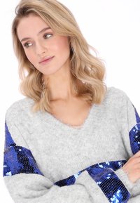 Grijze gebreide trui met wijde mouwen, V-hals en blauwe paillettenaccenten op de schouders. Zachte textuur, losse pasvorm en lichtgewicht ontwerp.