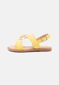 Sandalias amarillas con un upper de cuero suave, diseño abierto, correa de tobillo ajustable y adornos florales en la parte delantera. Suela de goma plana.
