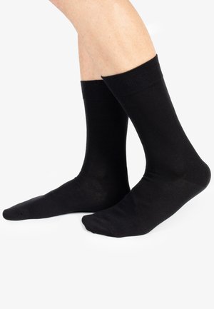 Calze di cotone nere con una texture liscia, bordino a coste e altezza alla caviglia, posizionate su piedi di tonalità della pelle chiara su uno sfondo bianco.