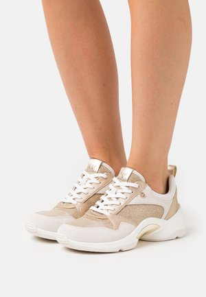 Piernas con zapatillas deportivas metálicas doradas y blancas con cordones y suelas blancas gruesas, de pie sobre un fondo claro liso.