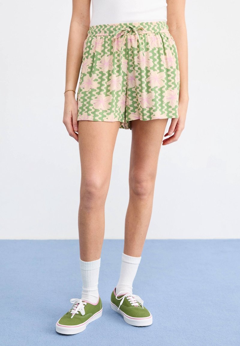Femme portant un short vert à motif floral, des chaussettes blanches et des baskets vertes, debout sur un sol bleu avec un fond blanc.