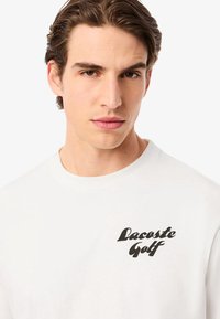 Camiseta de algodón blanca con un logo cursivo negro "Lacoste Golf" en el área del pecho izquierdo. Textura suave, diseño clásico de cuello redondo.