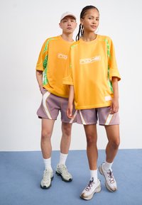 Două modele poartă ținute casual asortate, cu tricouri galbene supradimensionate ce au accente verzi și un logo, completate cu pantaloni scurți lila și pantofi sport.