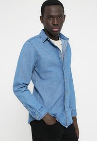 Világoskék farmerringgombos kialakítással, hegyes gallérral és hosszú ujjakkal, finom textúrával és normál szabással.