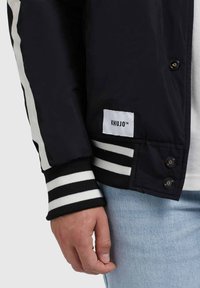 Zwarte jas met witte strepen, met knoopmanchetten en een merklabel. De stof lijkt glad en is gecombineerd met lichtblauwe jeans.