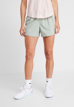 Frau trägt ein hellrosa ärmelloses Oberteil, hellgrüne Sportshorts, weiße Nike-Socken und weiße Laufschuhe, steht auf grauem Boden.