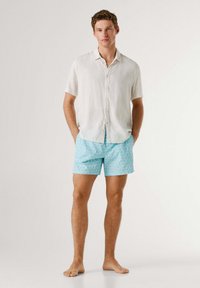 Ung mand står barfodet iført en hvid kortærmet skjorte med knapper og lyseblå shorts med hvidt geometrisk mønster.