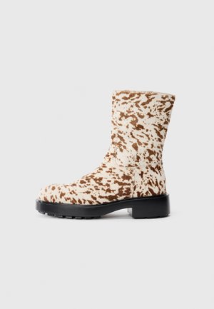Masivní kotníková bota vyrobená z texturovaného materiálu s krémovým základem a hnědým skvrnitým vzorem, vybavená černou gumovou podrážkou a kulatou špičkou.