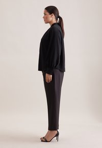 Blusa nera con maniche a palloncino e polsini con bottoni, abbinata a pantaloni neri sartoriali e sandali con tacco neri, vista di lato.