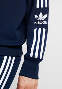 Sweatshirt bleu marine avec un grand logo Adidas blanc sur la manche, présentant trois bandes blanches le long des manches et un poignet côtelé.