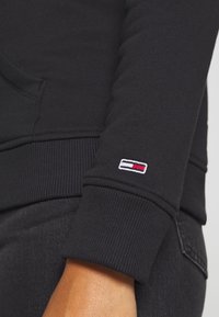 Acercamiento a la manga de un sudadera negra con puño acanalado y un pequeño logo rectangular rojo, blanco y azul cerca de la muñeca.