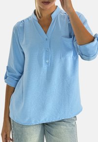 Hellblaues Hemd mit Knöpfen, Stehkragen, Brusttasche und hochgekrempelten Ärmeln aus strukturiertem Stoff. Getragen zu hellblauen Jeans.