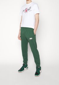 Vit t-shirt med svart och röd "Flight" grafik, gröna joggers med Nike-logga, och svarta/gröna sneakers med vita detaljer.